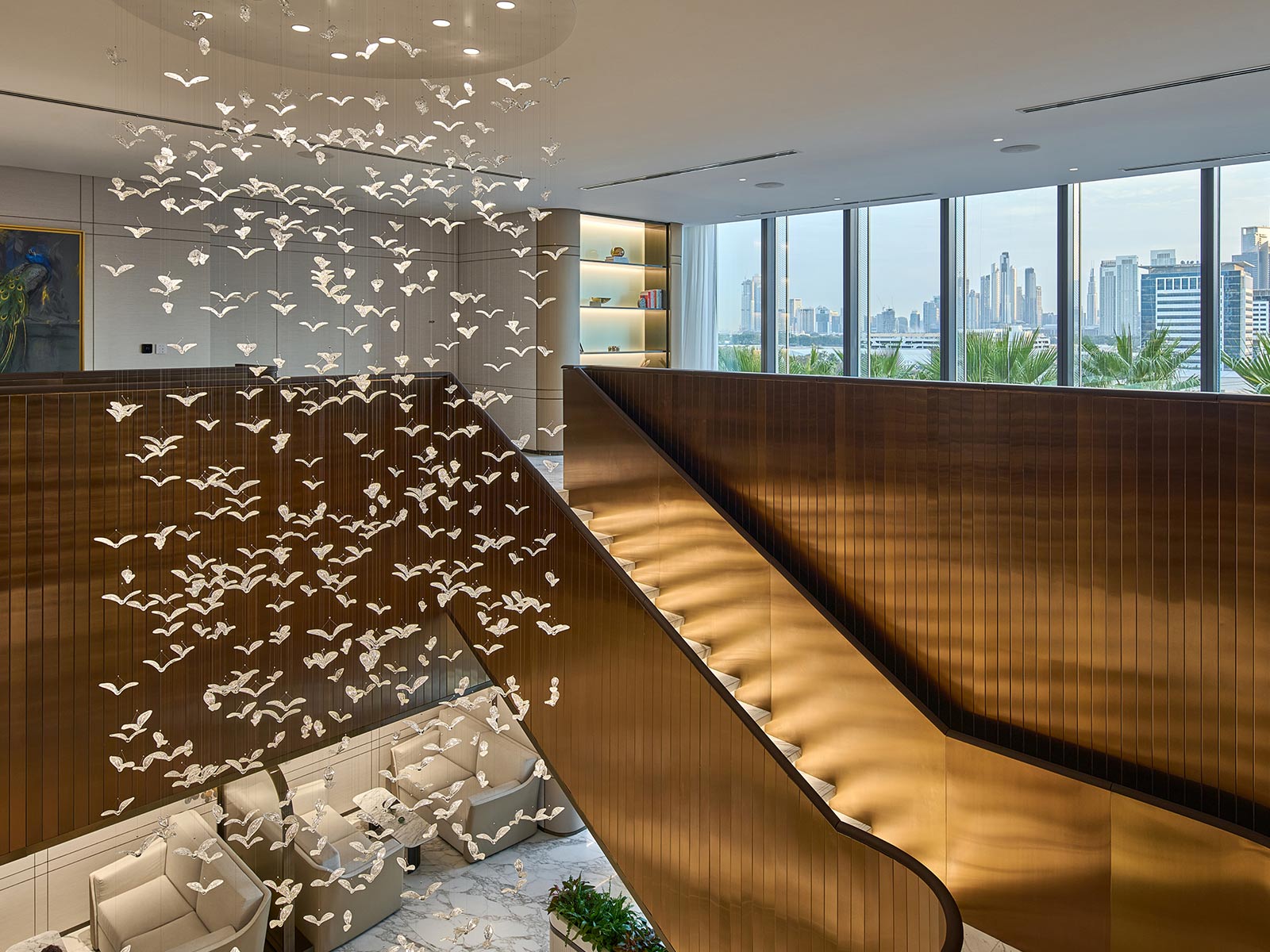 Handblown glass birds chandelier cascading above golden-hued office lobby staircase, by Nulty Bespoke